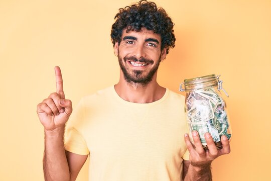 Handsome young man with curly hair and bear holding jar with savings smiling with an idea or question pointing finger with happy face, number one
