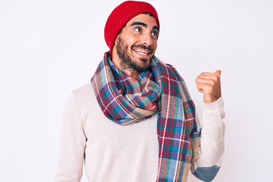 Handsome young man with curly hair and bear wearing a winter sweater, scarf and wool hat pointing thumb up to the side smiling happy with open mouth