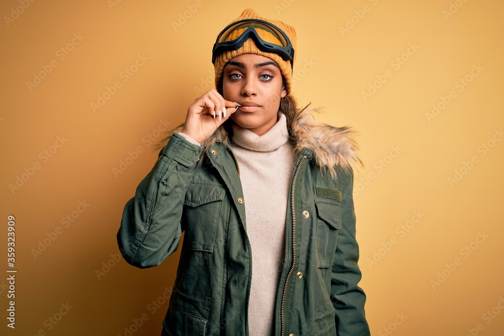 Wall mural Young african american afro skier girl wearing snow sportswear and ski goggles mouth and lips shut as zip with fingers. Secret and silent, taboo talking