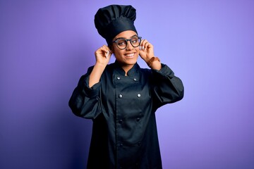 Young african american chef girl wearing cooker uniform and hat over purple background Smiling pulling ears with fingers, funny gesture. Audition problem