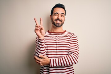 Young handsome man with beard wearing casual striped t-shirt standing over white background smiling with happy face winking at the camera doing victory sign. Number two.