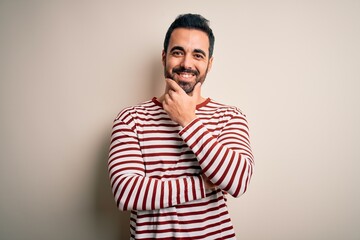 Young handsome man with beard wearing casual striped t-shirt standing over white background looking confident at the camera smiling with crossed arms and hand raised on chin. Thinking positive.