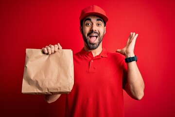 Young handsome delivery man with beard wearing cap holding takeaway paper bag with food very happy and excited, winner expression celebrating victory screaming with big smile and raised hands
