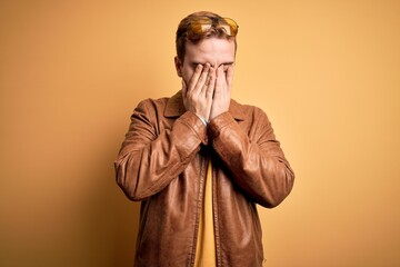 Young handsome redhead man wearing casual leather jacket over isolated yellow background with sad expression covering face with hands while crying. Depression concept.