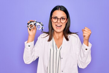 Young beautiful brunette oculist woman holding optometry glasses over purple background screaming proud, celebrating victory and success very excited with raised arms