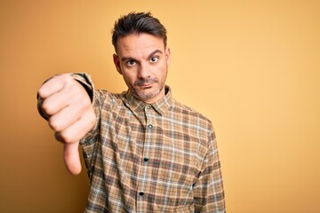 Young handsome man wearing casual shirt standing over isolated yellow background looking unhappy and angry showing rejection and negative with thumbs down gesture. Bad expression.