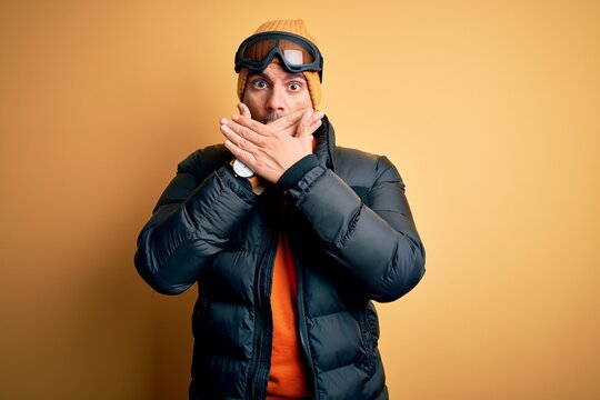 Young handsome skier man skiing wearing snow sportswear using ski goggles shocked covering mouth with hands for mistake. Secret concept.