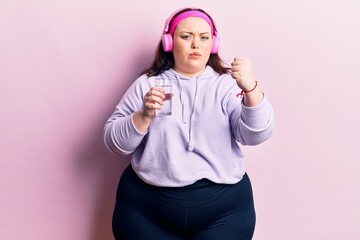 Obraz na płótnie Canvas Young plus size woman drinking glass of water using headphones annoyed and frustrated shouting with anger, yelling crazy with anger and hand raised