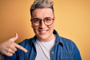 Close up of young handsome modern man wearing glasses and denim jacket over yellow background with surprise face pointing finger to himself