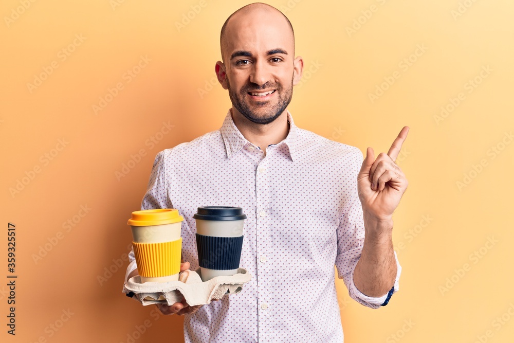 Wall mural young handsome bald man holding cup of takeaway coffee smiling happy pointing with hand and finger t