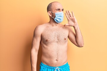 Fototapeta premium Young handsome bald man shirtless wearing swimwear and medical mask shouting and screaming loud to side with hand on mouth. communication concept.
