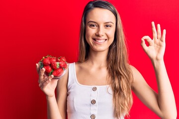 Young beautiful girl holding bowl with strawberries doing ok sign with fingers, smiling friendly gesturing excellent symbol