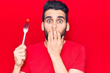 Young handsome man with beard holding fork with tomato covering mouth with hand, shocked and afraid...