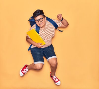 Adorable student boy wearing glasses and backpack smiling happy. Jumping with smile on face holding book celebrating with fist up over isolated yellow background.