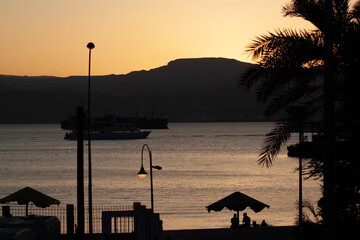 Sunset over the gulf of Aqaba. Silhouettes of trees, street lights and people under beach umbrellas are seen on the Jordanian coast while two ships are passing through the narrow strait. 