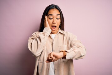 Young beautiful asian woman wearing casual shirt standing over pink background Looking at the watch time worried, afraid of getting late