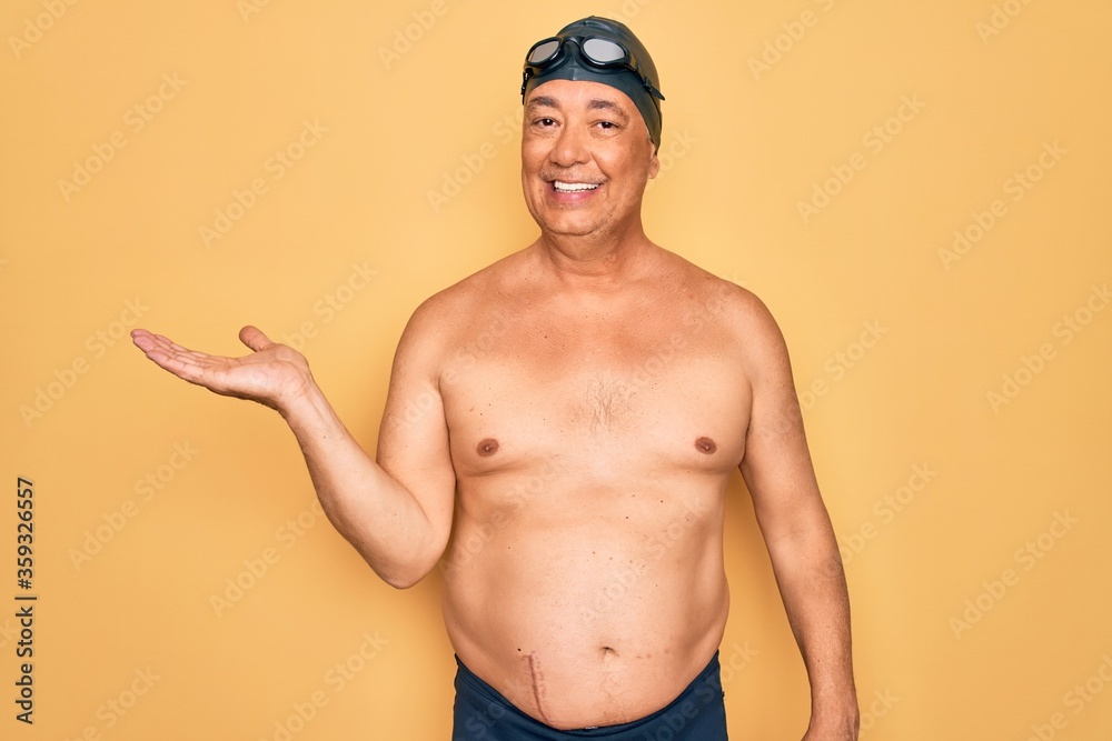 Canvas Prints Middle age senior grey-haired swimmer man wearing swimsuit, cap and goggles smiling cheerful presenting and pointing with palm of hand looking at the camera.