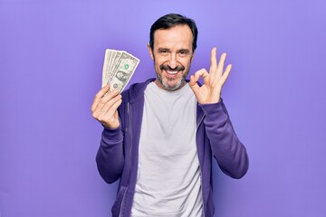 Middle age handsome man holding bunch of dollars banknotes over isolated purple background doing ok sign with fingers, smiling friendly gesturing excellent symbol