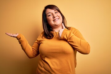 Beautiful brunette plus size woman wearing casual sweater over isolated yellow background Showing palm hand and doing ok gesture with thumbs up, smiling happy and cheerful
