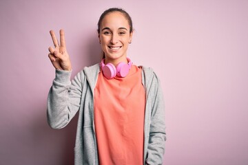 Beautiful sporty woman doing sport listening to music using headphones over pink background showing and pointing up with fingers number two while smiling confident and happy.