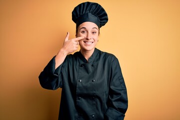 Young beautiful chef woman wearing cooker uniform and hat standing over yellow background Pointing with hand finger to face and nose, smiling cheerful. Beauty concept