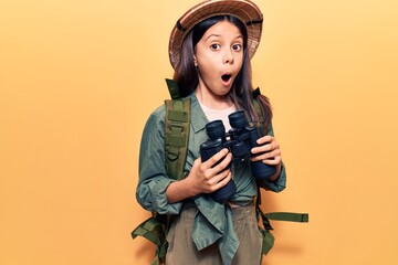 Beautiful child girl wearing explorer hat holding binoculars scared and amazed with open mouth for...