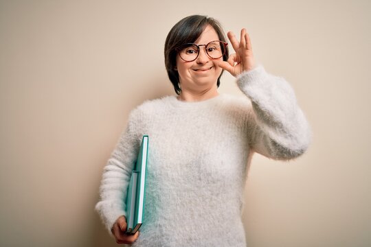 Young down syndrome student woman reading a book from library over isolated background doing ok sign with fingers, excellent symbol