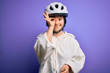 Young down syndrome cyclist woman wearing security bike helmet over purple background doing ok gesture with hand smiling, eye looking through fingers with happy face.