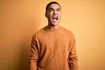 Young brazilian man wearing casual sweater standing over isolated yellow background angry and mad screaming frustrated and furious, shouting with anger. Rage and aggressive concept.