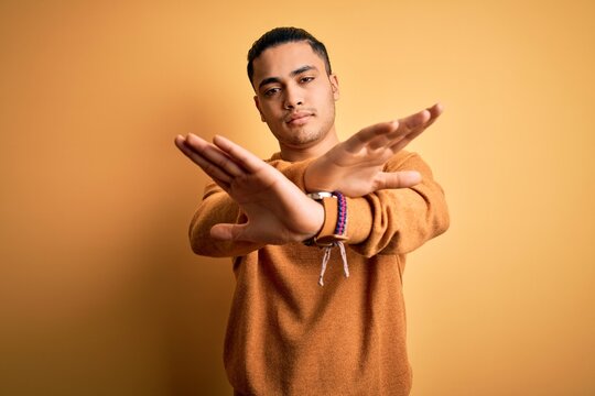 Young brazilian man wearing casual sweater standing over isolated yellow background Rejection expression crossing arms and palms doing negative sign, angry face
