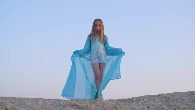 Adorable teenage girl in blue pareo standing on sand dune. Beautiful serious young girl in blue pareo dress standing on top of sandy hill and looking at camera