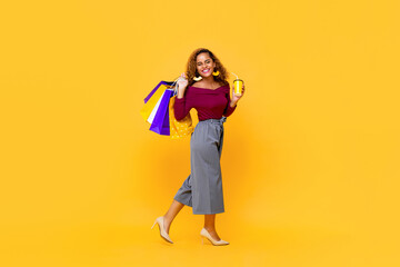 Fototapeta na wymiar Full length portrait of cheerful young smiling shopaholic African American woman walking while holding shopping bags and drink cup in isolated studio yellow background