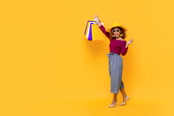 Full length portrait of smiling young attractive African American woman raising her shopping bags in isolated studio yellow background