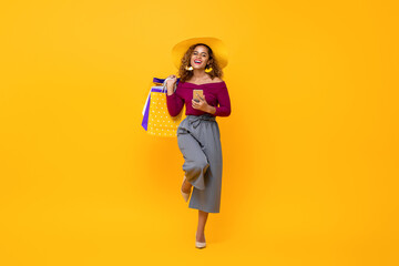 Full length portrait of happy young attractive African American woman holding shopping bags and mobile phone with one leg raised in isolated studio yellow background