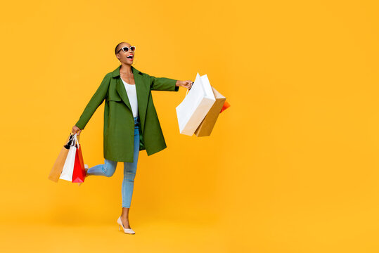 Full length portrait of cheerful attractive African American woman holding shopping bags while prancing in isolated studio yellow background
