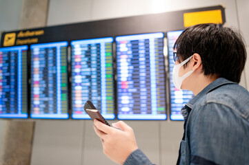 Asian man tourist wearing face mask checking flight from arrival departure board using smartphone in airport terminal. Coronavirus (COVID-19) pandemic prevention when travel. Social distancing concept