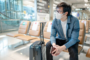 Asian man tourist wearing protective face mask sitting with suitcase luggage in airport terminal. Coronavirus (COVID-19) pandemic prevention when travel abroad. Health awareness and social distancing