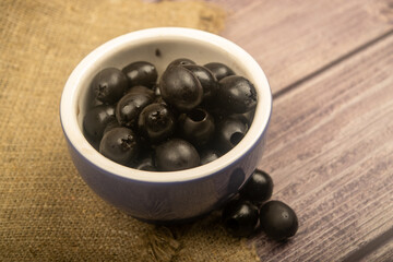 Black seedless olives in a ceramic bowl and a few scattered olives on a background of coarse-textured fabric. Close up.