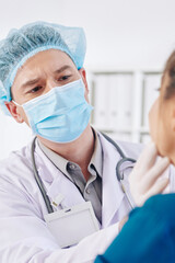 Serious general practitioner in medical mask examining glands and throat of female patient