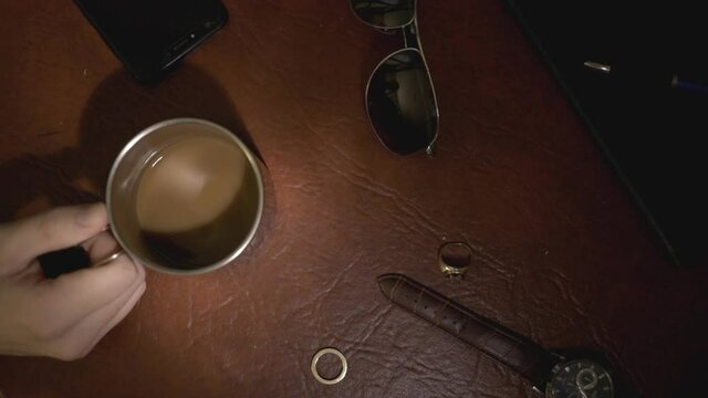 Mans Hand Holding A Cup Of Brown Coffee, Watch, Sunglasses, And A Pair Of Rings On The Brown Table - Flatlay - close up