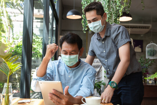 Portrait of a young man teaching his father how to use tablet computer and wearing mask for prevent the spread of the Covid-19. Father and Son using a tablet together at cafe.