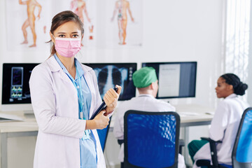 Young female doctor in medical mask holding tablet computer and looking at camera, her colleagues discussing x-rays and medical tests of patients in background