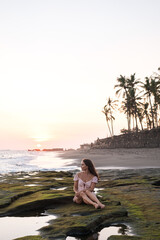Obraz premium Beautiful young girl in a dress sits on stones on the ocean at sunset. A girl in full growth looks into the distance at the ocean.