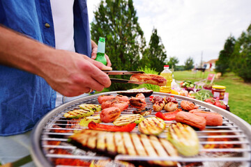 close-up of a man's hand holding a barbecue tongs with a delicious fried sausage on the background of a barbecue grill with sausages, vegetables, baked zucchini and pepper, beer bottles