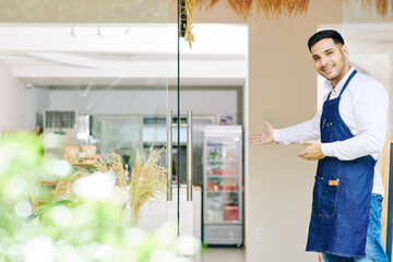 Fototapeta na wymiar Smiling young Indian man in denim apron inviting customers in bakery he just opened