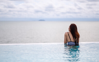 A young asian woman relaxing in infinity swimming pool looking at a beautiful sea view