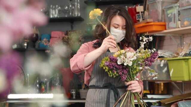 female adding new flower to bouquet. asian woman florist holding a half made arrangement and adding plants to composition. wearing mask due to flu. Designing, floral workshop, working concept