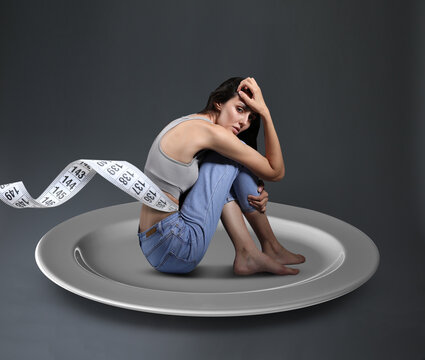 Small woman with measuring tape sitting on plate against grey background. Concept of anorexia