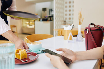 Waiter putting cup of coffee and delicious dessert on cafe table in front of customer