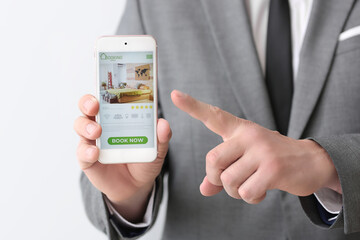 Businessman with mobile phone booking room in hotel online against light background